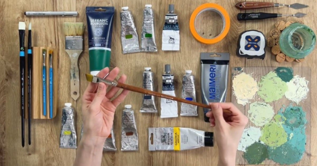 Acrylic painting masterclass art supplies on a wooden table seen from overhead with a pair of hands holding a brush